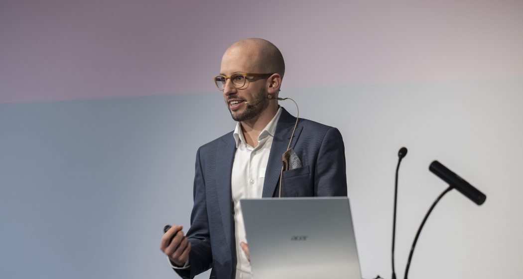 Marcel Meyer steht auf einer Bühne hinter einem Rednerpult und spricht mit Headset-Mikrofon vor Publikum. Vor ihm steht ein offener Laptop.