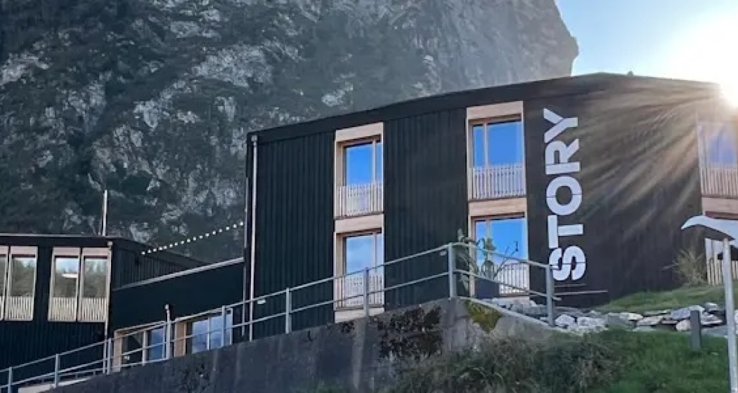 Aussenansicht des Story Thusis vor Bergkulisse bei Sonnenschein, mit moderner dunkler Holzfassade und vertikalem «STORY»-Schriftzug am Gebäude.