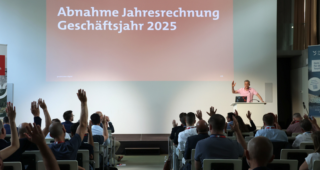 Abstimmung an der Generalversammlung von graubünden digital 2026: Die Teilnehmenden heben die Hand zur Annahme der Jahresrechnung 2025, während dJon Erni am Rednerpult auf der Bühne ebenfalls die Hand hebt. Im Hintergrund ist die Präsentationsfolie mit der Aufschrift «Abnahme Jahresrechnung Geschäftsjahr 2025» zu sehen.