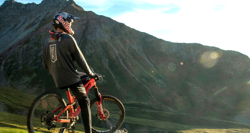 Mountainbiker mit Red Bull Helm und Bike Kingdom Trikot steht mit seinem Bike auf einer Alp, Bergpanorama im Hintergrund.