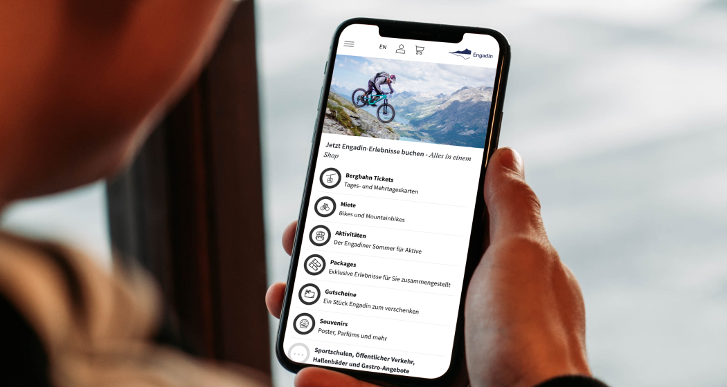 Person hält Smartphone in der Hand und nutzt die Engadin-App mit Buchungsoptionen für Bergbahn-Tickets, Bike-Miete und Aktivitäten, im Hintergrund Bergpanorama.