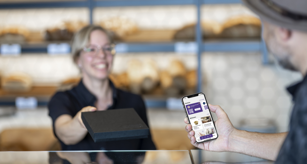 Bäckerei-Verkäuferin reicht einem Kunden über die Theke eine schwarze Schachtel. Der Kunde hält ein Smartphone mit geöffneter App (Bereiche «Vereine» und «Aktionen»).