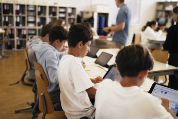 Schülerinnen und Schüler arbeiten im Klassenzimmer mit Tablets, während eine Lehrperson im Hintergrund unterrichtet