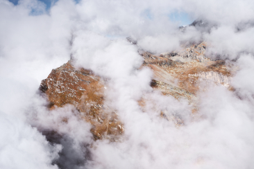 Felsige Berggipfel der Giandas, halb in Wolken gehüllt. Die rotbraunen Felswände ragen dramatisch aus dem Wolkenmeer empor, darüber blauer Himmel. Foto: Gaudenz Danuser.