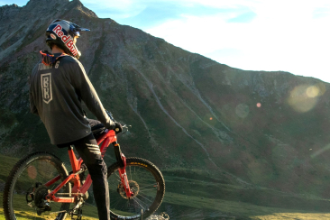 Mountainbiker mit Red Bull Helm und Bike Kingdom Trikot steht mit seinem Bike auf einer Alp, Bergpanorama im Hintergrund.